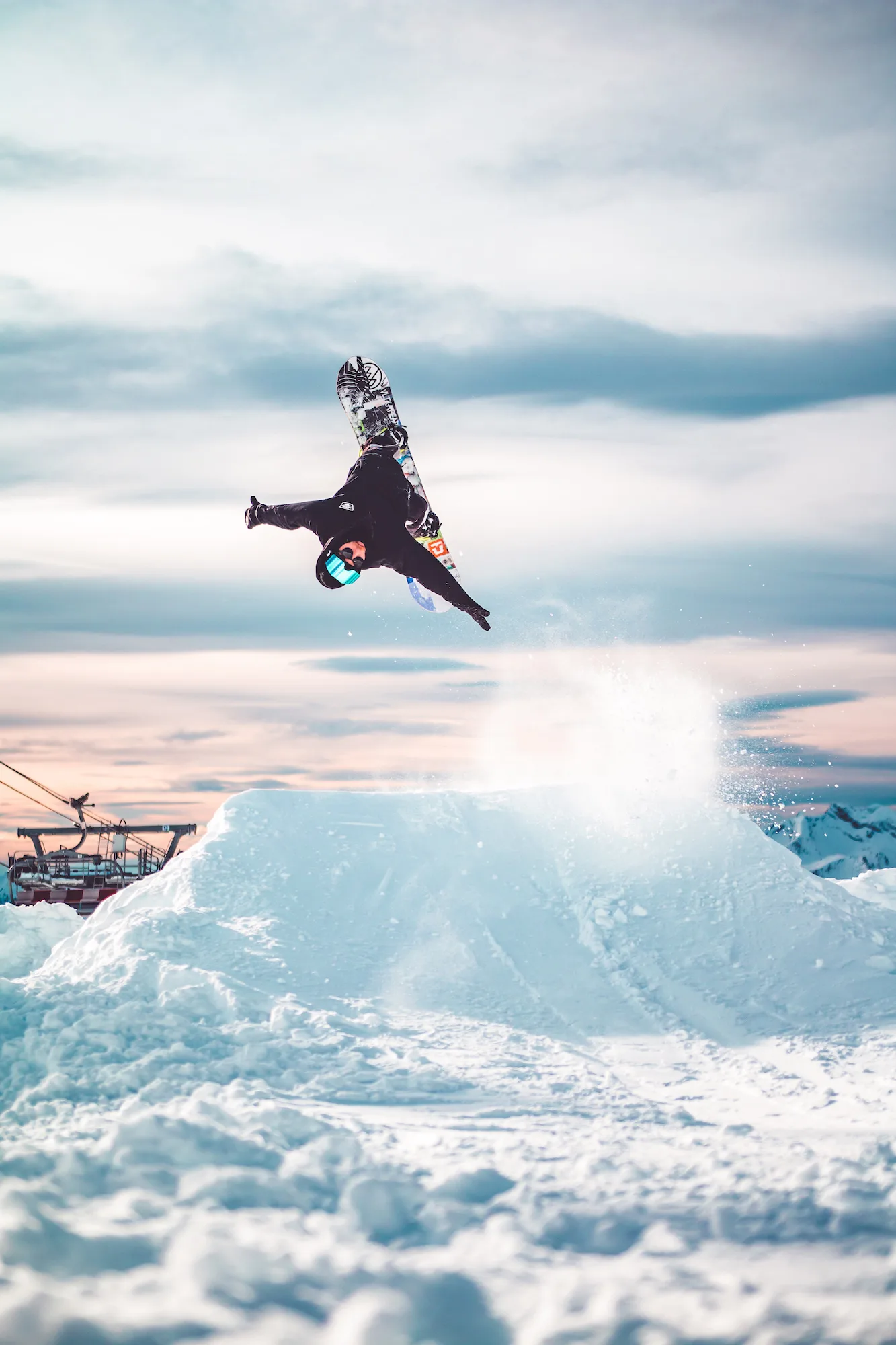 Snowboarder upside down in mid-air above a snow jump at sunset, with a chairlift in the background