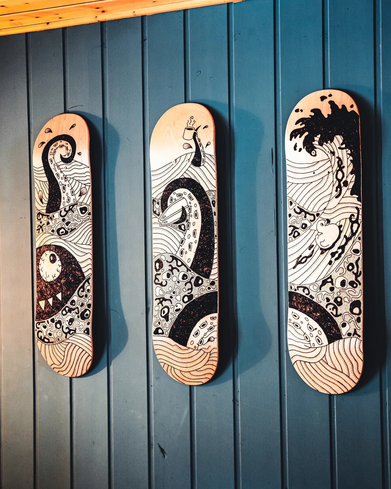Three skateboard decks with hand-drawn tentacle, eyeball and wave artwork on a blue wooden wall
