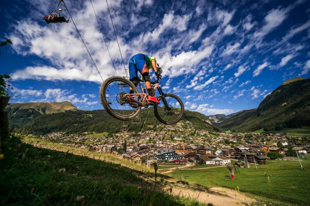Mountain biker jumping through the air beside a chairlift line, with Morzine village in the valley below