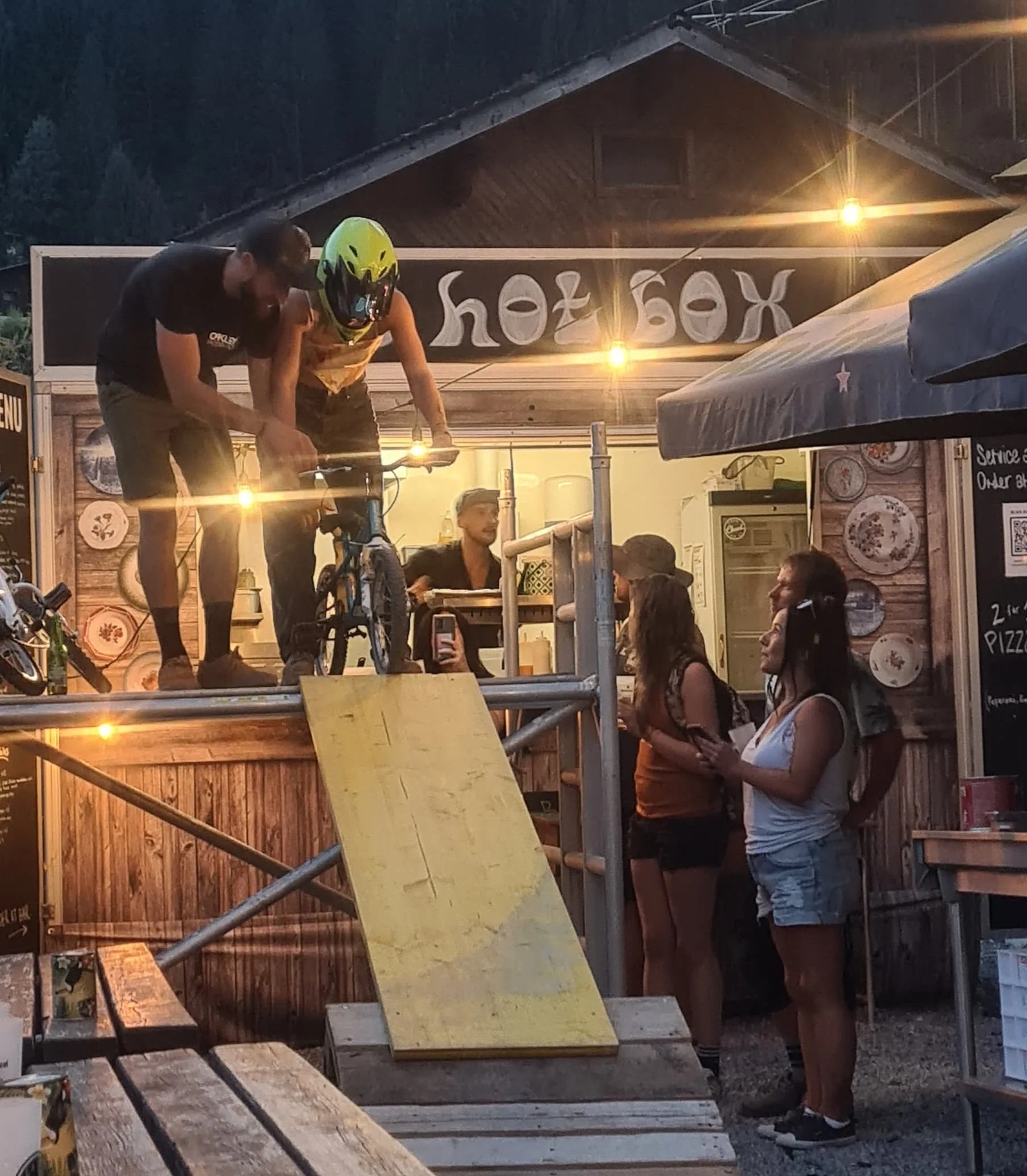 Fahrer mit grünem Helm oben auf einer Holzrampe vor dem Hot Box Food-Hüttchen, beobachtet von Zuschauern
