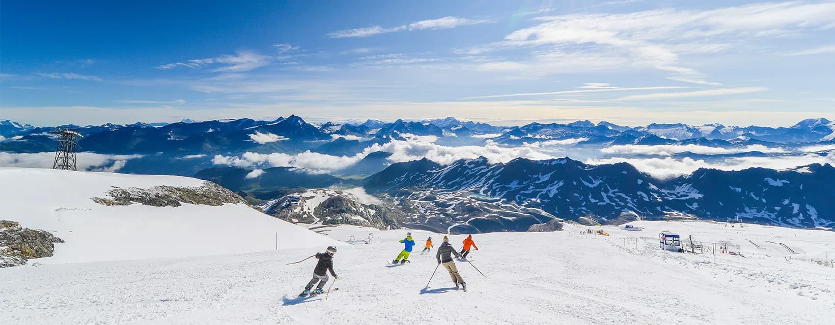 Gruppe von Skifahrern fährt einen sonnigen Hang hinunter, dahinter ein weites Bergpanorama