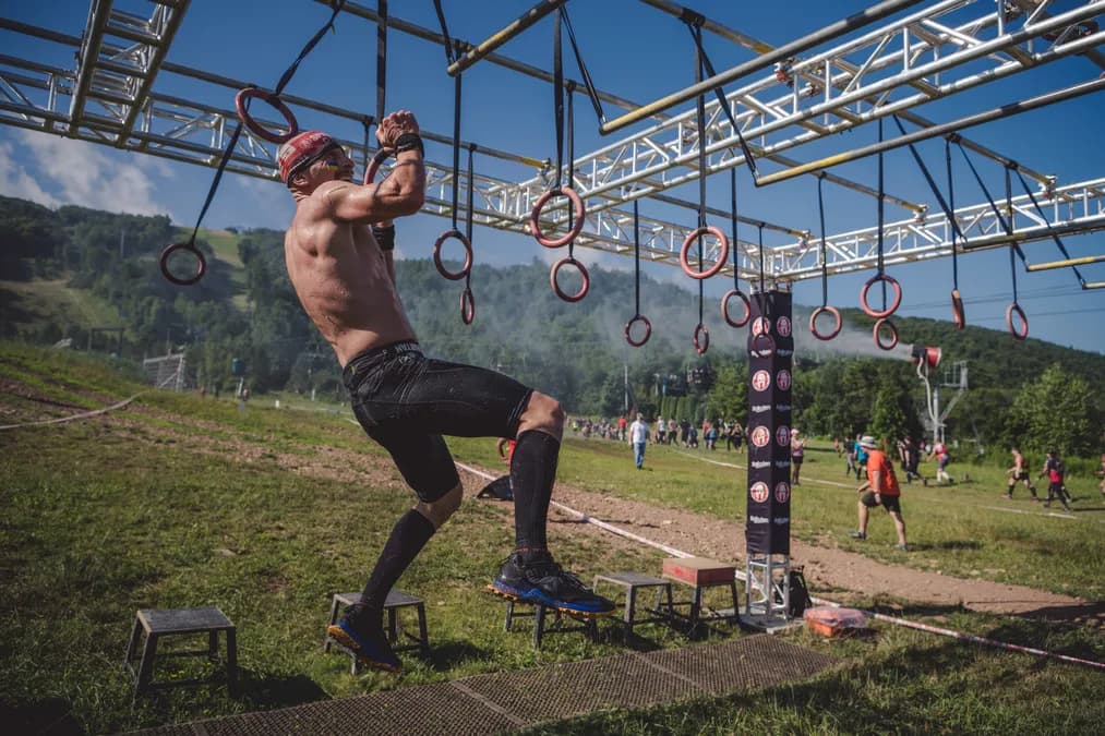 Mann schwingt sich bei einem Spartan Race an hängenden Ringen über ein Hindernis auf einer Bergwiese