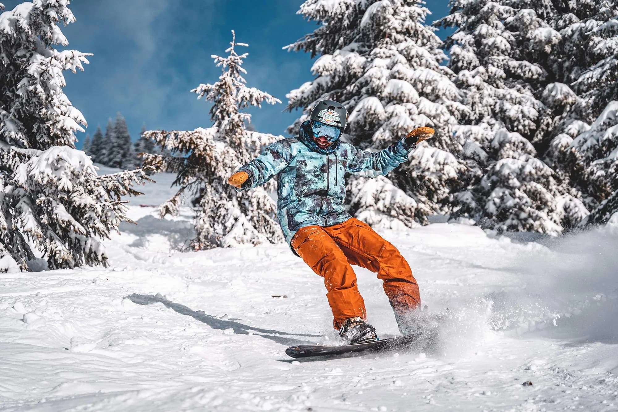 Snowboarder in orangefarbener Hose carvt durch Pulverschnee zwischen schneebedeckten Tannen