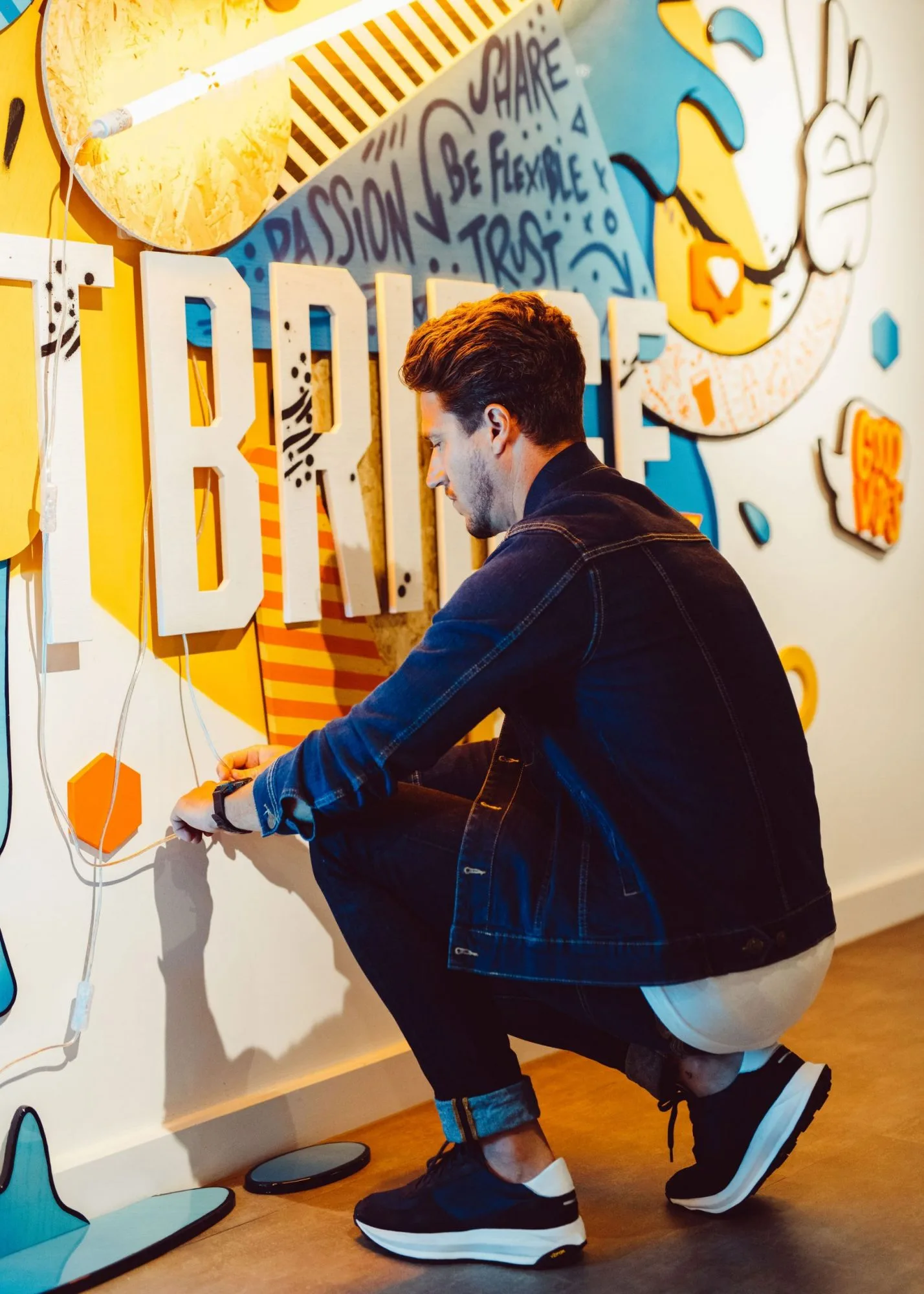 Mann in Jeansjacke hockt neben einem bunten Wandbild mit Buchstaben und richtet ein Kabel