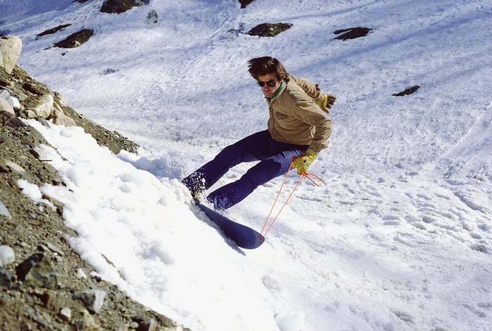 Oude foto van een skiër in beige jas en zonnebril die langs rotsen een besneeuwde helling af zwaait