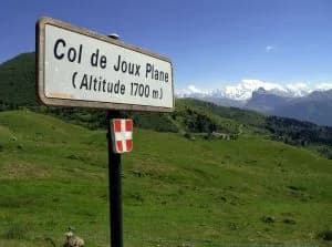 Sign reading “Col de Joux Plane (Altitude 1700 m)” with green meadows and snowy peaks behind