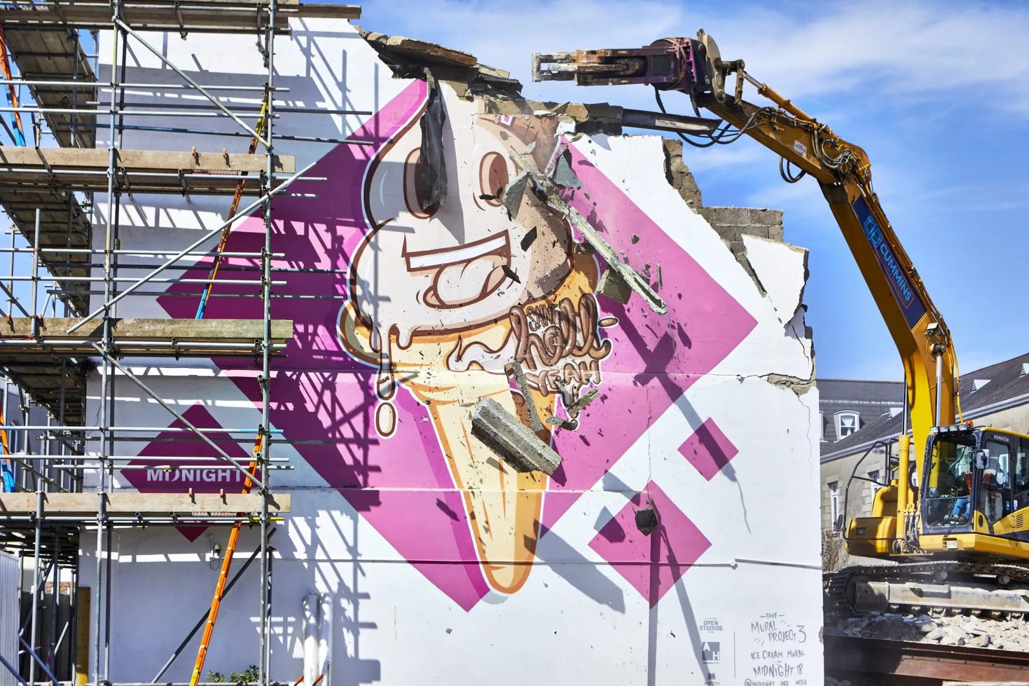 Ice cream cone mural on a building wall being demolished by an excavator, with scaffolding alongside