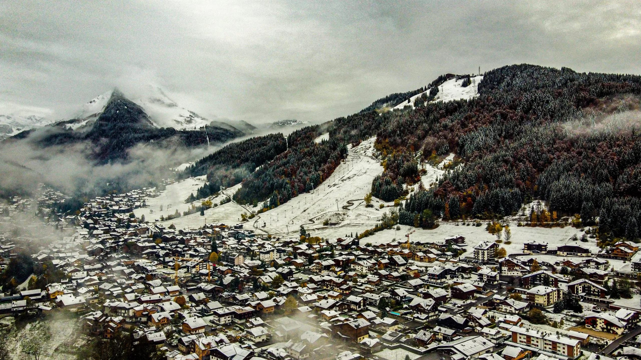 Luftaufnahme der Dächer von Morzine unter leichtem Schnee, dahinter bewaldete Hänge und neblige Gipfel
