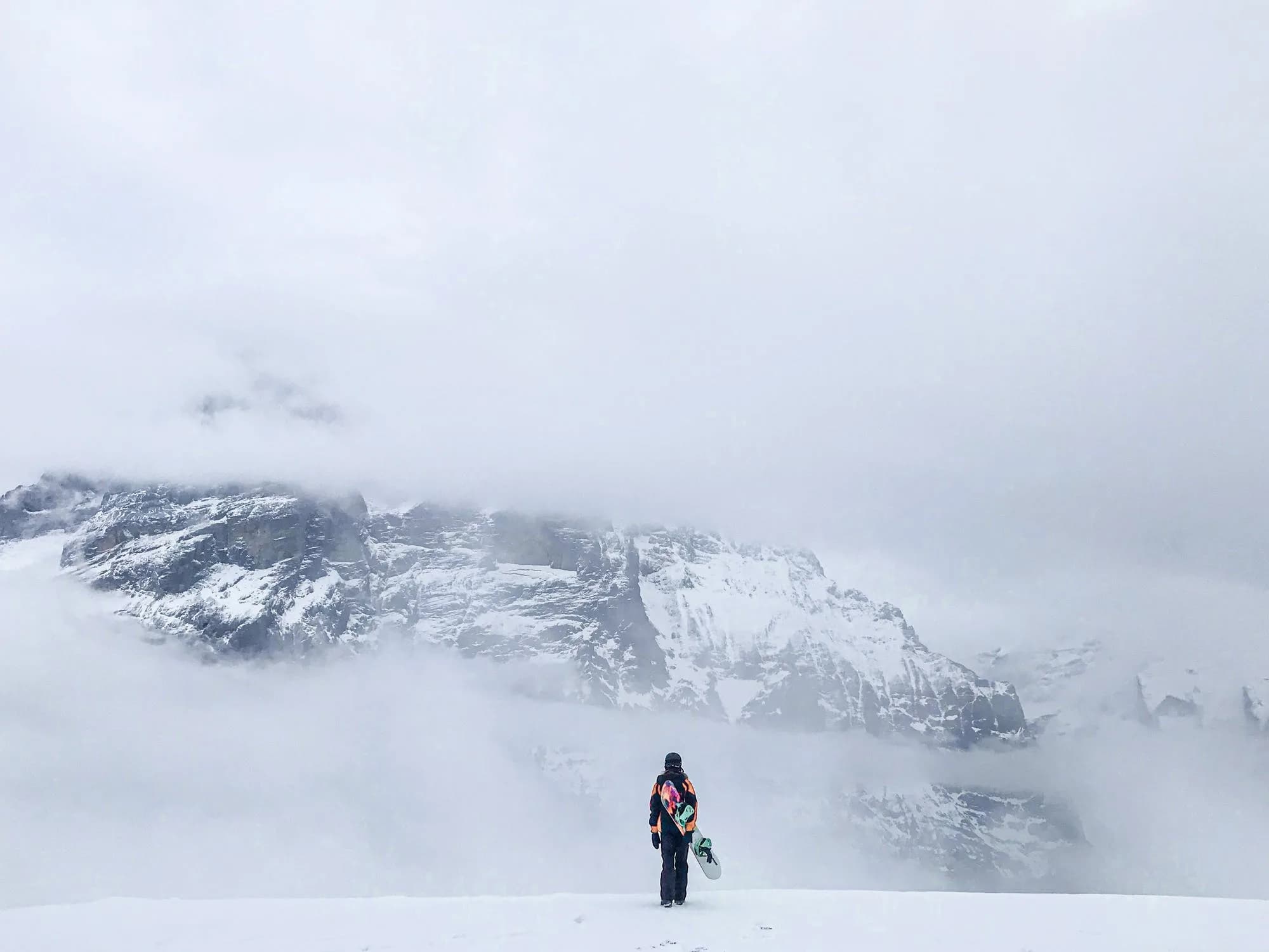 Snowboarderin trägt ihr Board über ein verschneites Plateau zu wolkenverhangenen Bergen