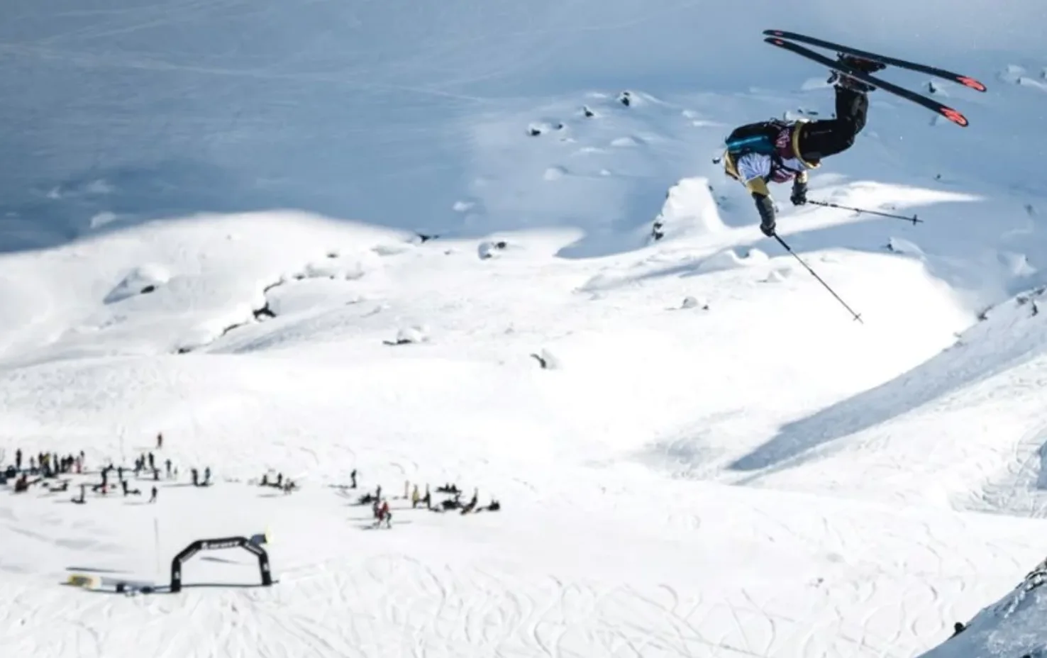 Skiër maakt een achterwaartse salto hoog boven een besneeuwde helling, met toeschouwers en een finishboog beneden