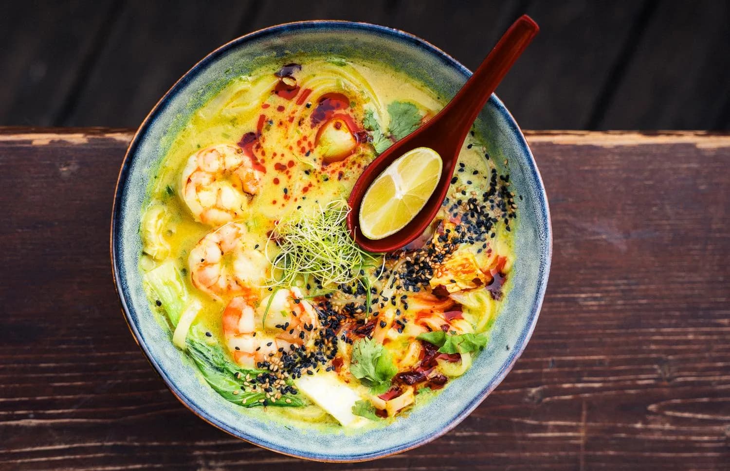 Eine goldgelbe Nudelbowl mit Garnelen, Grünzeug und einem Löffel voller Limette